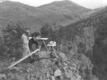 Three men perched on a peak with a plane table that supports several instruments. One man is writing in a notebook, another is peering through an optical survey instrument. Another is poised at a desk writing.